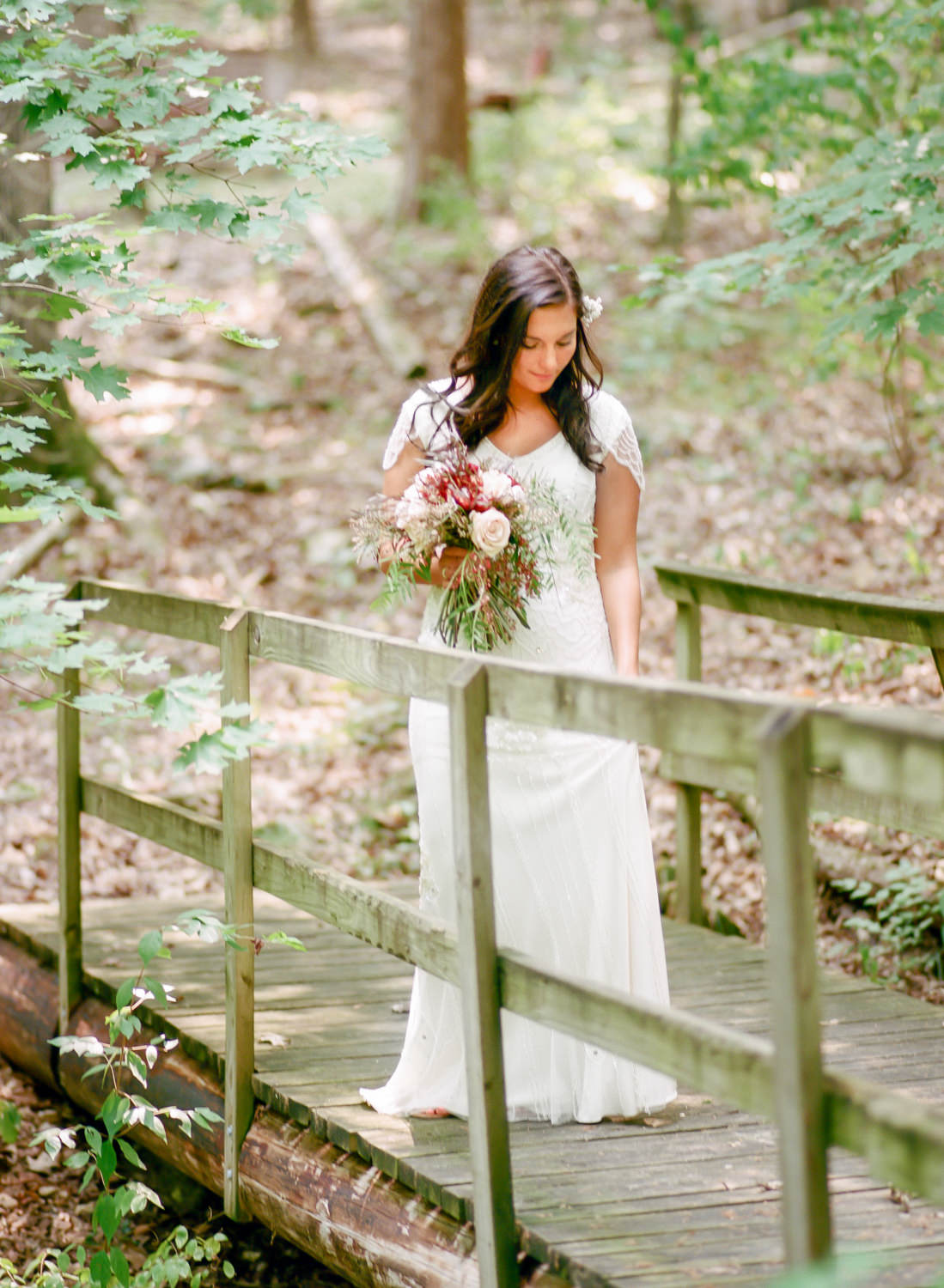 bride in woods on bridge in beaded gown and holding rustic red bouquet, Erica Robnett Photography